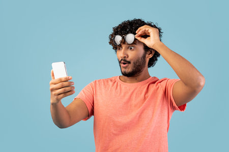 Shocked Eastern Man With Curly Hair Looks at Smartphone in Blue Studio Background