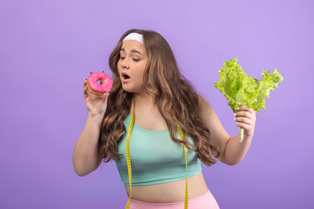 A Funny Young European Woman in Sportswear Holds a Donut in One Hand and a Salad in the Otherの写真素材