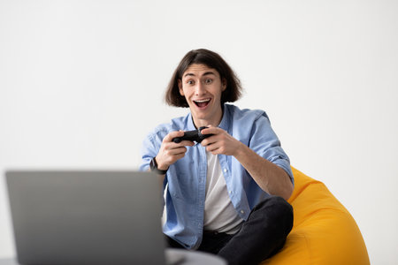 Young Man Enjoys Gaming on a Laptop While Sitting on a Bean Bag in a Bright Roomの写真素材