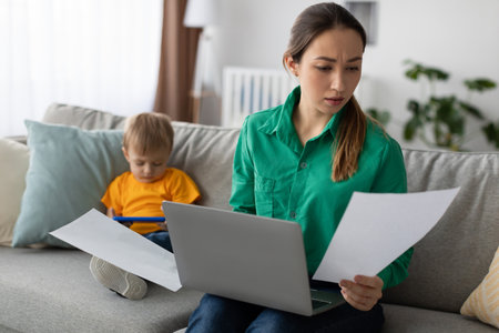 Busy Mother Working on Tasks While Supervising Her Child at Home in a Cozy Living Roomの写真素材