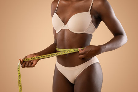 Young Woman Measuring Her Waist Using Tape in a Beige Studio Settingの写真素材