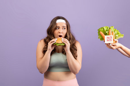 Hungry Young Lady in Sports Uniform Enjoys Burger While Receiving Vegetables on Purple Backgroundの写真素材