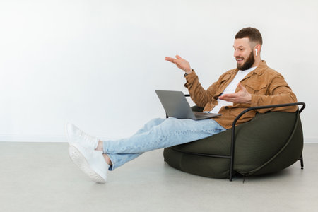 Man Smiling and Talking While Sitting in a Bean Bag Chair Using a Laptop During a Casual Online Meetingの写真素材