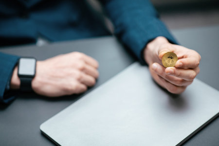 Person Holds a Gold Coin and Sits at a Desk With a Laptop, Showcasing Interest in Cryptocurrency in a Modern Office Settingの写真素材