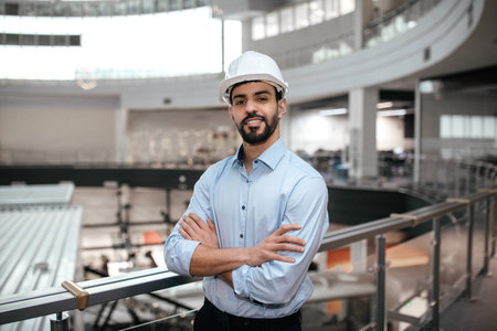 Young Engineer Stands Confidently in a Modern Industrial Facility With Arms Crossedの写真素材