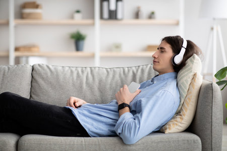 Young Man Relaxing on Couch Listening to Music With Headphones at Home in a Cozy Environmentの写真素材