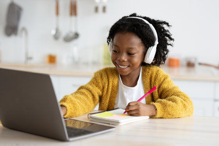 Joyful Child Engages With Online Learning While Taking Notes in Cozy Kitchen Settingの写真素材