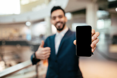 Businessman Shows Smartphone With Blank Screen While Giving Thumbs up in Modern Office Spaceの写真素材