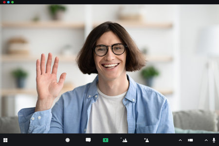 Young Man Smiles and Waves During a Virtual Meeting at Home in Comfortable Settingの写真素材