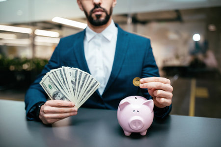 Businessman Holding Cash and Inserting Coin in Pink Piggy Bank in Modern Office Settingの写真素材