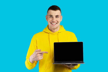Young Man in Yellow Hoodie Holding a Laptop and Smiling Against a Bright Blue Background