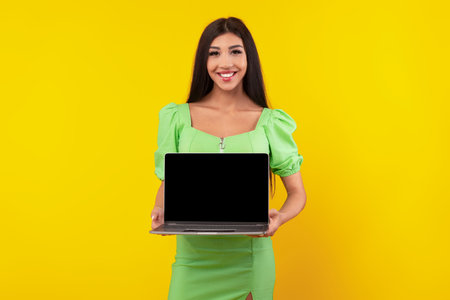 Woman in Green Dress Holding Laptop Against Bright Yellow Background, Showcasing Technology With a Smileの写真素材