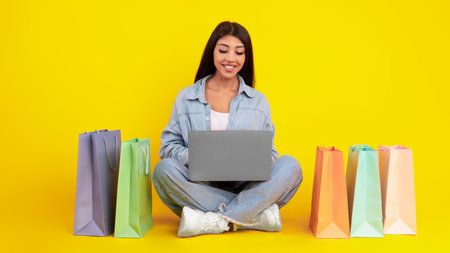 Young Woman Shopping Online With a Laptop and Colorful Bags Against a Vibrant Yellow Backgroundの写真素材