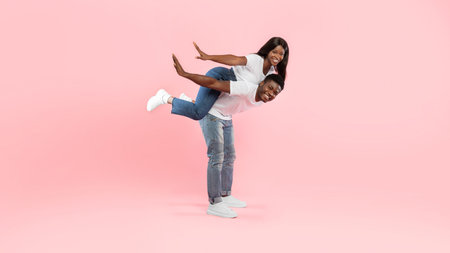 Smiling Black Man Carries His Woman on His Back in Playful Moment Against a Pink Background