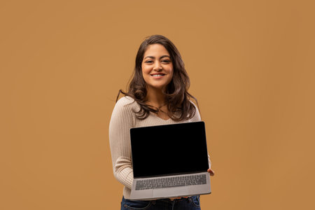 Happy arab lady showing laptop with blank screen and smiling to camera, standing over beige background, mockupの写真素材