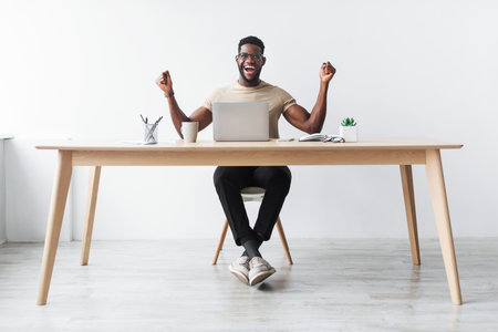 Young Black Office Employee Celebrates Success at Desk With Laptop and Joyful Energyの写真素材