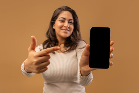 Mobile app concept. Cheerful arab woman pointing at smartphone with blank screen over brown backgroundの写真素材