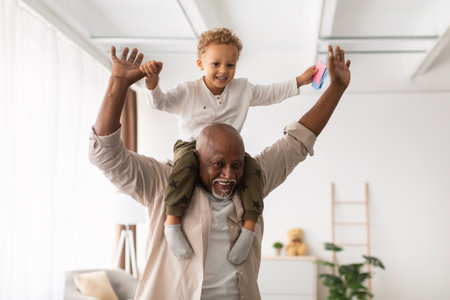 Cheerful Black Grandpa Carrying Smiling Grandson on Shoulders Indoors During Weekend Playtimeの写真素材