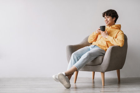 Young Asian Man Enjoys Coffee While Relaxing in Armchair During Peaceful Morningの写真素材