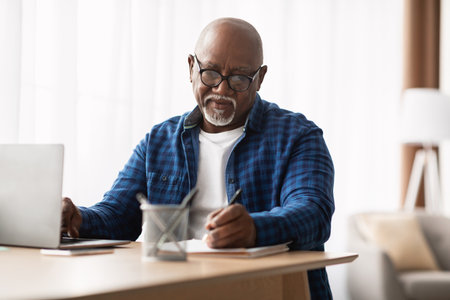 Mature African American Man Taking Notes While Using Laptop in Modern Office Environmentの写真素材