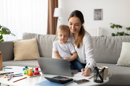 Mother Works on Laptop While Child Plays and Colors at Home During the Dayの写真素材