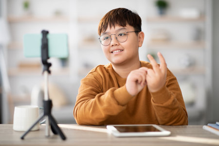 Cute Asian Teen Boy Vlogger Streams From Home Using Cellphone and Tablet on a Tripodの写真素材