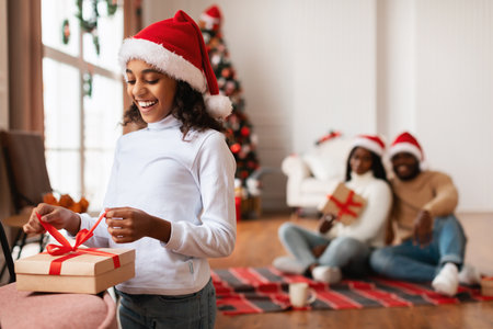 Excited Black Girl Unwraps Christmas Gift With Family Celebrating in Living Roomの写真素材