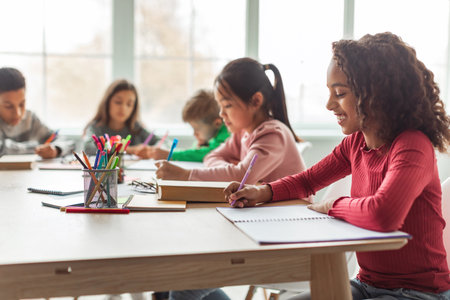 African American School Girl Learns and Writes at Desk in Classroom With Other Childrenの写真素材
