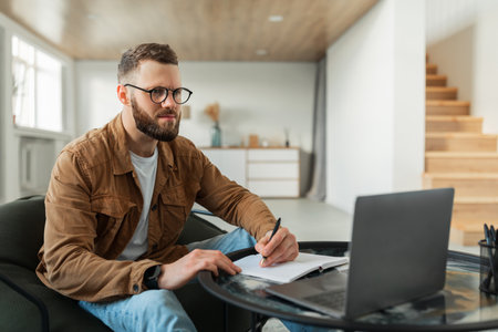 Focused Man Writes Notes While Working on Laptop in a Cozy Modern Living Room Settingの写真素材