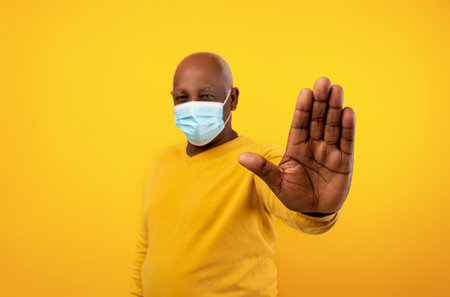 Senior Black Man Gesturing Stop in Face Mask Against Orange Background During Coronavirus Pandemicの写真素材