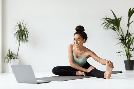 Beautiful Young Woman Enjoys Stretching Leg Muscles at Home While Using a Laptop for Workoutsの写真素材