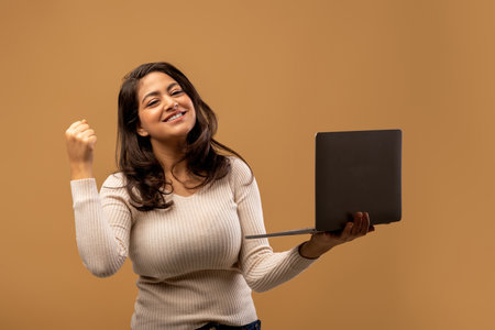 Work online at home. Happy arab lady holding laptop and expressing emotions of success, brown background, studio shotの写真素材