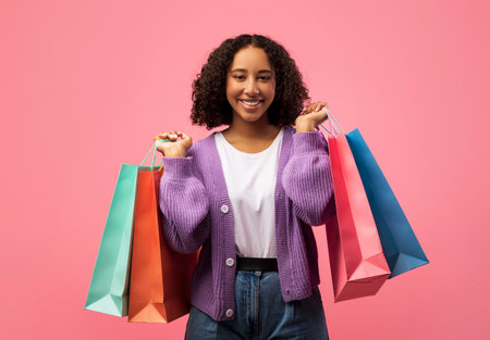Charming Young Black Woman Enjoys Shopping With Colorful Bags on Pink Backdropの写真素材