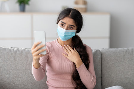 Young Indian Woman in Mask Makes Video Call on Sofa During Covid-19 Quarantineの写真素材