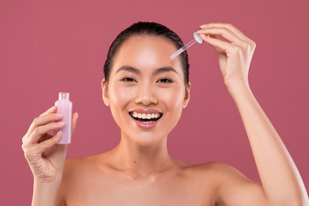Beautiful Young Woman Smiling While Applying Face Serum Against a Purple Studio Backgroundの写真素材