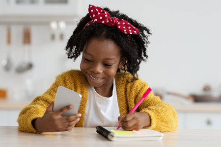 Young Girl With Curly Hair Smiles While Using Smartphone and Writing in Notebook at Homeの写真素材