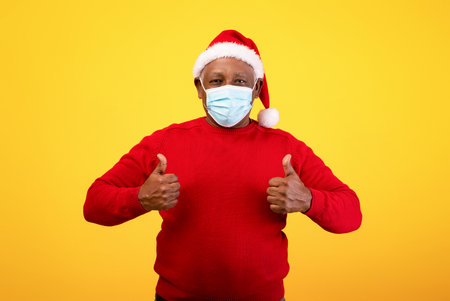 Senior Black Man in Santa Hat and Face Mask Gives Thumbs up for Christmas Celebrationの写真素材
