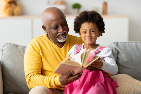 Smiling Girl and Grandfather Enjoying Fairy Tales on the Sofa During Cozy Evening at Homeの写真素材