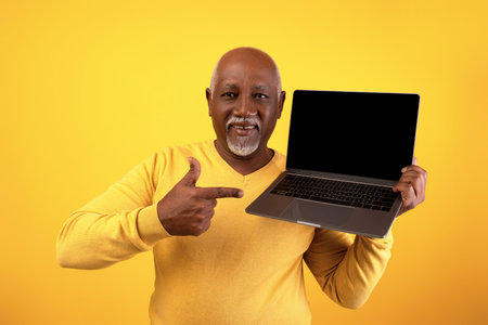 Positive Elderly Black Man Points at Laptop on Orange Background for Website Mockupの写真素材