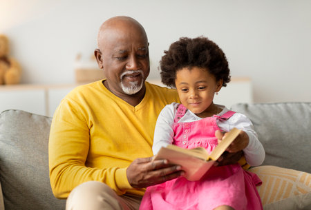 Elderly Man and Little Girl Enjoy Reading Fairy Tales on a Cozy Sofa in the Living Roomの写真素材