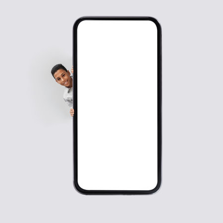 Black Boy Smiles While Posing Near Large Smartphone With Blank Screen on a Gray Backgroundの写真素材