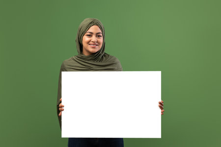 Happy Muslim Lady With Empty Placard for Advertisement Over Dark Green Backgroundの写真素材