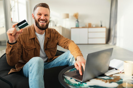 Man Smiling While Holding a Credit Card and Using a Laptop in a Modern Living Room Setting During Daylight Hoursの写真素材