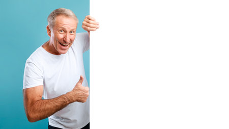 Mature Man Shows Thumbs up While Peeking From Behind a Blank Advertising Boardの写真素材