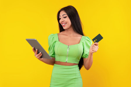 Woman in Green Outfit Using Tablet and Holding Credit Card With Yellow Background While Smiling and Enjoying Online Shopping Experienceの写真素材