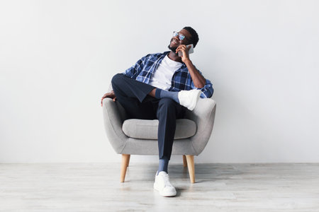 Young Black Man Enjoys a Cheerful Phone Conversation While Sitting in an Armchairの写真素材