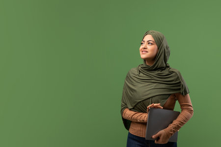 Cheerful Arab Lady in Hijab Holding Laptop and Looking Aside Against Green Backgroundの写真素材