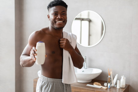 Smiling Black Male Holds Shower Gel Bottle in Home Bathroom While Wrapped in Towelの写真素材