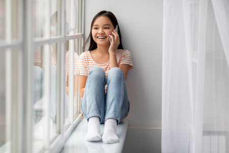 Young Girl Sitting on a Windowsill Talking on the Phone in a Bright, Cozy Room During the Dayの写真素材