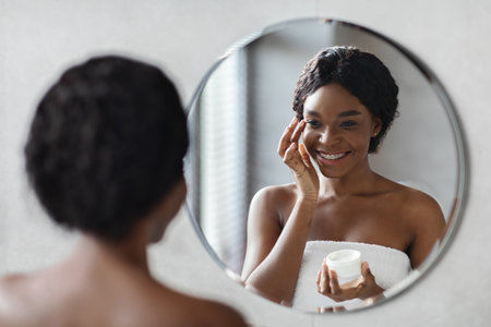 Happy Young Black Woman Applying Cream in Mirror After Morning Shower for Face Careの写真素材